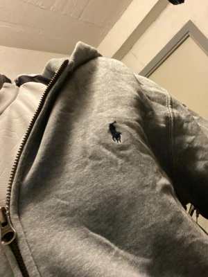 Grå zip hoodie från Polo Ralph Lauren - Säljer en grå hoodie med dragkedja från Polo Ralph Lauren. Klassisk design med mörkblå logga broderad på bröstet, två fickor framtill och huva. Tillverkad i mjukt sweatshirtmaterial som är skönt att ha på sig.