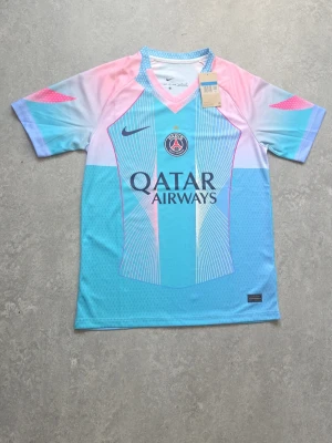 PSG x Nike pastell fotbollströja - En riktigt snygg PSG fotbollströja i pastellfärger från Nike. Tröjan har ett coolt mönster i ljusblått, rosa och vitt, med PSG-logga och Qatar Airways tryck på bröstet. Perfekt för sommaren och det varma vädret.