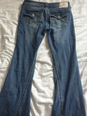 Blå bootcut jeans med broderade fickor - Säljer ett par blå bootcut jeans med snygga broderade bakfickor och dekorativa knappar. Jeansen har klassisk femficksdesign och är gjorda i denim med lätt tvättad look. Perfekta för dig som gillar retrostil och vill ha ett par jeans med unik detalj på fickorna.