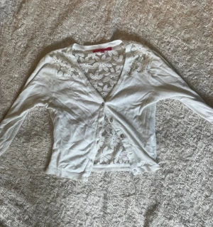 Vit kofta med spetsdetaljer Derhy Kids - Söt vit kofta från Derhy Kids med genomskinliga spetsdetaljer och blommönster på ryggen och axlarna. Koftan har v-ringning, knäppning framtill och ribbade muddar. Perfekt för att lyfta en outfit med en romantisk touch.