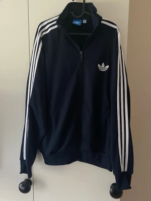  Adidas track jacka - Snygg marinblå track jacka från Adidas med klassiska vita ränder längs ärmarna och dragkedja framtill. Tröjan har hög krage, ribbade muddar och Adidas logga broderad på bröstet. Perfekt för en sportig och avslappnad stil.