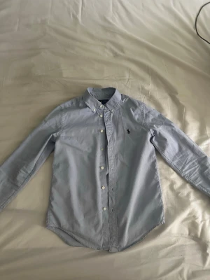 Ljusblå skjorta från Ralph Lauren - Klassisk ljusblå skjorta från Ralph Lauren med button-down krage och broderad logga på bröstet. Skjortan har långa ärmar, vita knappar och är tillverkad i bomull. Perfekt för en clean och stilren look.❗️Pris kan sänkas❗️