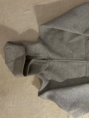 Grå zip hoodie med huva - En stilren grå hoodie med dragkedja framtill och huva. Tillverkad i mjukt sweatshirtmaterial och har långa ärmar samt fickor på sidorna. Perfekt för en avslappnad look och enkel att matcha med andra plagg.