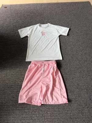 T-shirt - Säljer ett par ljusrosa shorts och en vit T-shirt med elastisk midja och vit snörning. Shortsen har en avslappnad passform och är tillverkade i ett mjukt syntetmaterial. Broderad textdetalj på ena sidan ger en extra touch. Perfekta för sommardagar eller chill hemma.