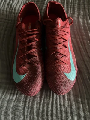 Röda Nike fotbollsskor med turkos swoosh - Säljer ett par röda Nike fotbollsskor med turkos swoosh på sidan. Skorna är i relativt bra skick men har använts en del men har fortfarande mycket mer i sig. Skriv vid funderingar och priset är inte hugget i sten och kan rabatteras vid snabbt köp och köp av flera annonser!
