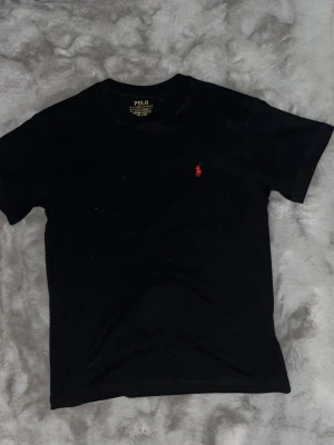 Svart t-shirt från Polo Ralph Lauren - Helt oanvänd svart t-shirt från Polo Ralph Lauren med röd broderad logga på bröstet. Modellen är slim fit och tillverkad i 100% bomull. Perfekt för en stilren och klassisk look.