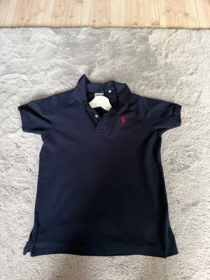 Marinblå piké t-shirt med röd polo assn  - Klassisk marinblå piké t-shirt med krage och knappar framtill. T-shirten har en röd broderad pollo aspen logga på bröstet och är gjord i mjuk bomull. Passformen är normal och ärmarna är korta, perfekt för en clean och enkel stil.