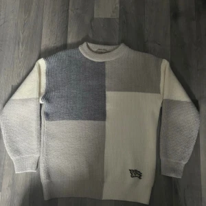 Blockmönstrad stickad burberry tröja i grått och vitt - Säljer en stickad burberry tröja med blockmönster i grått och vitt. Tröjan har rund hals, ribbade muddar och en liten broderad detalj nertill. Perfekt för dig som gillar en clean men ändå unik look. Materialet känns mjukt och tjockt, perfekt för kyligare dagar.