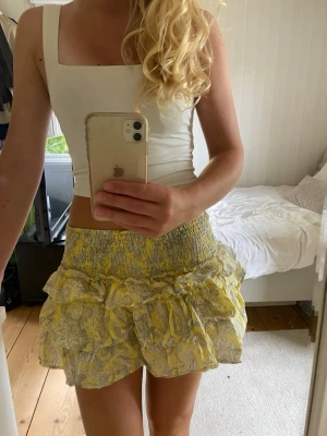 Gul volangkjol med blommönster - Söt kort kjol i gult med volanger och blommigt mönster. Kjolen har bred smock i midjan för skön passform och är gjord i ett lätt bomullstyg. Perfekt för varma dagar och ger en lekfull vibe till din outfit.