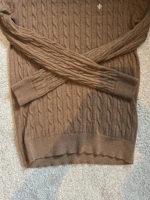 Kabelstickad Morris tröja - Snygg brun kabelstickad tröja med rund hals och långa ärmar. Nästan aldrig använd och den är som nyskick. Tröjan har ribbade muddar vid ärmslut och nederkant samt ett diskret broderat emblem på bröstet. 
