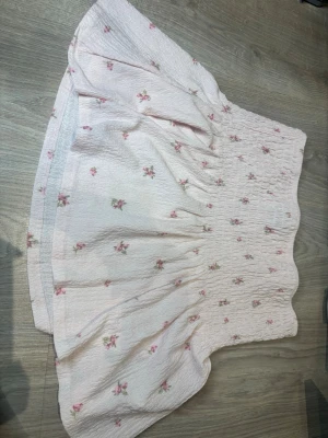 Blommig kortkjol  - Söt kortkjol från Gina Tricot i ljusrosa med små blommiga mönster i rosa och grönt. Kjolen har en smockad midja och volangkant nedtill, vilket ger en romantisk och somrig känsla. Perfekt för dig som gillar en feminin och trendig stil. Oanvänd