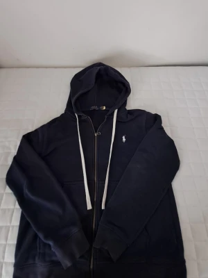 Marinblå hoodie från Polo Ralph Lauren - Snygg marinblå hoodie från Polo Ralph Lauren med dragkedja framtill, huva med vita snören och ribbade muddar. Tillverkad i mjukt bomullsmaterial som känns skönt mot huden. Perfekt för en avslappnad och stilren look.