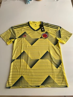 Helt ny! Colombianske 2019 hemma - Snygg gul matchtröja från Adidas med Colombias landslagsmärke på bröstet. Tröjan har blåa och röda detaljer, V-ringning och korta ärmar. Mönstret består av blåa diagonala linjer och trianglar som ger en sportig look. Perfekt för fotboll och träning.