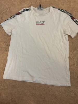 Vit EA7 Emporio Armani t-shirt - Säljer en vit t-shirt från EA7 Emporio Armani med logga på bröstet och svarta band med text på axlarna. Klassisk rund hals och korta ärmar. Perfekt för dig som gillar stilrena märkesplagg.