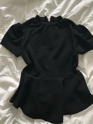 Svart peplum topp med puffärm - Snygg svart topp med peplumskärning och puffiga korta ärmar. Hög halsringning och mjukt, följsamt material som ger en stilren look. Perfekt att styla med jeans eller kjol för en trendig outfit.
