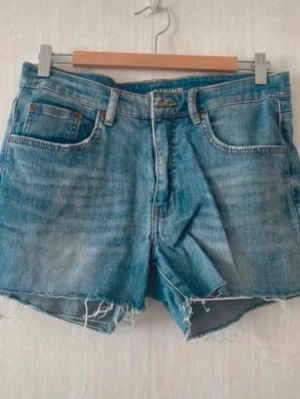 Blå denimshorts med rå kant - Säljer ett par klassiska blå denimshorts i storlek 38. Shortsen har fem fickor, normal passform och rå, fransig kant nedtill för en avslappnad look. Perfekta till sommaren och gjorda i slitstark bomull.