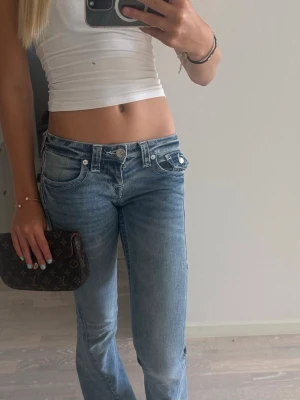 Glittriga True religion jeans - Säljer ett par ljusblå jeans med låg midja och bootcut-modell. Jeansen har klassiska fem fickor, snygga slitningar och en avslappnad passform. Perfekta för dig som gillar en trendig och avslappnad stil. JOEY 