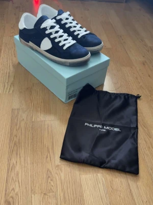 Philippe Model blåvita sneakers - Snygga sneakers från Philippe Model i marinblått och vitt. Skorna har en klassisk låg siluett, vita skosnören och detaljer i mocka och läder. Kommer med originalpåse och låda. Perfekta för dig som gillar stilrena och trendiga skor.