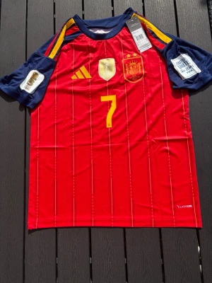Spaniens matchtröja Ferran nr 7 - Röd och marinblå matchtröja från Adidas för Spaniens landslag, med gula detaljer och nummer 7 på både fram- och baksida. Namnet 'Ferran' står på ryggen. Tröjan har officiella märken och CLIMACOOL-material för bästa komfort under match.