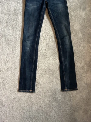 Mörkblå Nudie jeans 25/32 - Snygga mörkblå skinny jeans med slitningar och klassiska orange sömmar. Jeansen har en smal passform och normal midja, perfekta till en avslappnad stil. Materialet är denim med lite stretch för extra komfort.