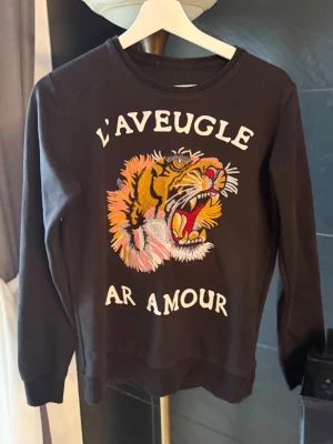 Svart sweatshirt med tigerbrodyr - Cool svart sweatshirt med stor broderad tiger och texten 'L'AVEUGLE PAR AMOUR' på framsidan. Tröjan har rund halsringning och långa ärmar. Materialet ser ut att vara mjukt och bekvämt, perfekt för en avslappnad stil.