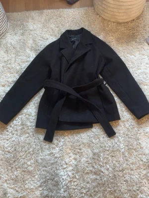 Svart kappa från Vero Moda med skärp - Stilren svart kappa från Vero Moda med klassisk krage och knytbart midjebälte. Kappan har rak passform och är helfodrad för extra komfort. Perfekt för dig som vill ha en enkel men snygg look till hösten eller vintern.
