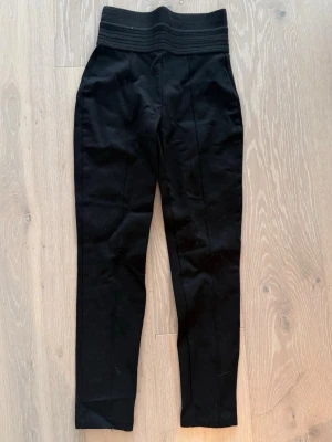 Svarta kostymbyxor med hög midja - Stilrena svarta kostymbyxor med hög midja och bred resår i midjan. Byxorna har raka ben och pressveck framtill för en snygg och modern look. Perfekta till en dressad outfit och passar till många olika stilar.