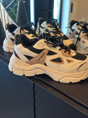 Chunky sneakers från Steve Madden - Chunky sneakers i beige, vitt och svart från Steve Madden. Skorna har meshpaneler för extra ventilation, detaljer i syntetläder och en rejäl sula som ger streetwear-vibbar. Svarta snören och kontrasterande färgblock gör dem till ett statement i varje outfit.