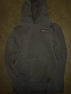 Mörkblå hoodie från Peak Performance - Snygg mörkblå hoodie från Peak Performance med klassisk logga på bröstet. Hoodien har stor huva och en rymlig magficka framtill. Perfekt för dig som gillar en enkel och stilren look. Tillverkad i mjukt material för extra komfort.
