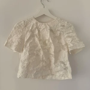 Vit blommig kortärmad blus - Säljer en söt vit blus med blommigt mönster i strukturen. Toppen har rund halsringning och korta ärmar. Perfekt för dig som vill ha en fräsch och stilren look med lite extra detalj. Passar till både jeans och kjol!