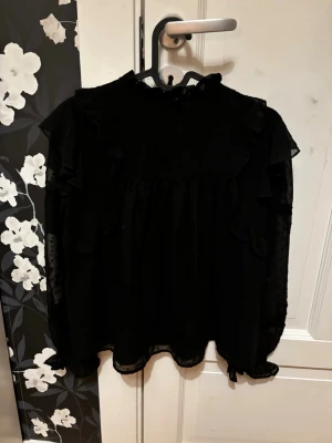 Svart volangblus med transparenta ärmar - Snygg svart blus med volangdetaljer och hög krage. Ärmarna är långa och transparenta med diskreta prickar och spetsdetaljer. Blusen har en luftig passform och är gjord i ett tunt, skönt material som ger en elegant look.