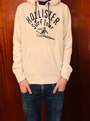 Vit hoodie från Hollister med tryck - Säljer en vit hoodie från Hollister med svart tryck på bröstet där det står 'Hollister Surf Tour' och en surfare. Hoodien har huva med snören och en stor magficka. Perfekt för dig som gillar en avslappnad och sportig stil.