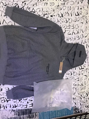 Grå Essentials Fear of God hoodie - Säljer en grå hoodie från Essentials Fear of God med svart tryck på bröstet och ryggen. Hoodien har en klassisk huva, ribbade muddar och magficka. Perfekt för dig som gillar streetwear och vill ha en stilren look.