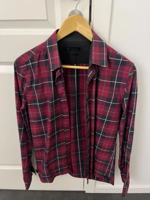 Rutig röd skjorta från Riley - Säljer en snygg röd och svart rutig skjorta från Riley i slim fit-modell. Skjortan har långa ärmar, klassisk krage och knappar framtill. Perfekt för dig som gillar en stilren men ändå avslappnad look.