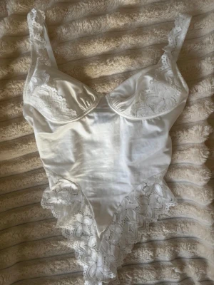 Vit satinbody med spetsdetaljer - Supersnygg vit body i satin med spetsdetaljer på kupor, axelband och längs höfterna. Bodyn har bygelkupor och tunna axelband i spets för en extra feminin look. Materialet är glansigt och känns lyxigt mot huden. Perfekt för dig som vill ha något elegant och romantiskt.