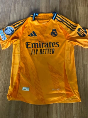 Orange  Real Madrid fotbollströja Adidas - Snygg orange Real Madrid matchtröja från Adidas med mörkblå detaljer och tryck. Tröjan har klubbmärke, sponsorlogga och nummer 9 med Mbappé på ryggen. Tillverkad i lätt och ventilerande material, perfekt för fotbollsträning eller match.