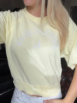 Ljusgul kortärmad topp med tryck - Säljer en ljusgul topp med texten 'Malibu California LA' i vitt framtill. Toppen har korta ärmar, rund halsringning och är gjord i mjuk bomull. Perfekt för dig som gillar en avslappnad och fräsch stil.💙