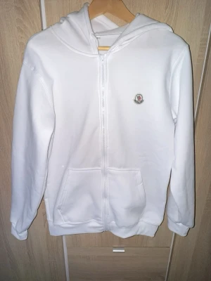 Moncler hoodie - Säljer en stilren vit hoodie med dragkedja framtill och praktisk huva. Hoodien har två stora fickor fram och en liten logga på bröstet. Perfekt för dig som gillar enkel och clean stil. Materialet känns mjukt och bekvämt.