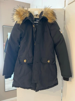 Svart parkasjacka med fuskpäls från Svea - Snygg svart parkasjacka från Svea med avtagbar fuskpäls på huvan och två stora fickor framtill. Jackan har dragkedja och knappar, samt beige innerfoder. Materialet är 60% bomull och 40% nylon, fyllning 80% dun och 20% fjäder.