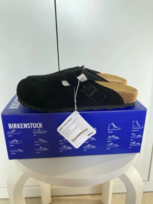 Svarta Birkenstock Boston tofflor - Säljer ett par svarta Birkenstock Boston tofflor i mocka med justerbart spänne och mjuk fotbädd. Klassisk slip-in modell med korksula och greppvänlig yttersula. Perfekta för chill dagar hemma eller när du vill ha det bekvämt.