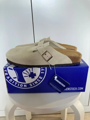 Beige Birkenstock tofflor mocka - Snygga beige tofflor från Birkenstock i mjuk mocka med klassisk design och justerbart spänne i metall. Fotbädden är i kork och innersulan i mocka för extra komfort. Perfekta för dig som vill ha både stil och bekvämlighet.