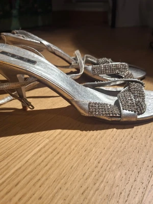 Silvriga sandaletter med strassdetaljer - Säljer ett par glittriga sandaletter i silver med smala remmar och dekorerade med strass framtill. Klackarna är smala och ger en elegant look. Perfekta för dig som vill sticka ut och addera lite extra bling till outfiten. Materialet är syntet med metallic-finish.