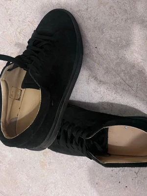 Svarta låga sneakers i mocka - Stilrena svarta sneakers i mocka med lågt skaft och svart sula. Skorna har klassisk snörning och en enkel, clean design som passar till det mesta. Insidan är ljus och kontrasterar snyggt mot utsidan.