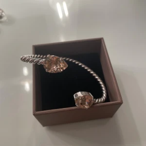 Carolina Svedbom Silvrigt armband med ljusrosa stenar - Snyggt armband i silverfärgad metall med tvinnad design och två stora ljusrosa droppformade stenar i varje ände. Stenarna har en glittrig finish som fångar ljuset och ger extra bling. Perfekt för dig som gillar statement-smycken med unik form.