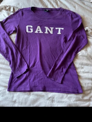Lila långärmad topp från GANT - Lila långärmad topp från GANT med vit logotyp framtill. Tillverkad i mjuk bomull och har en klassisk rund halsringning. Perfekt för en avslappnad och stilren look.