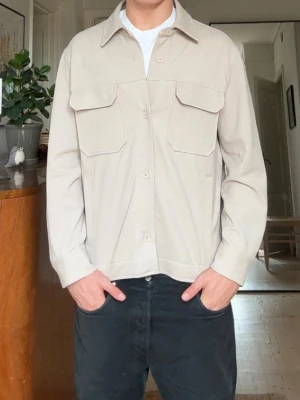 Beige overshirt med stora fickor - Stilren beige overshirt med knäppning framtill och två stora bröstfickor med lock. Rak passform och klassisk krage. Tillverkad i ett mjukt, vävt material som ger en avslappnad look. Perfekt att bära över en t-shirt för en enkel och snygg stil.