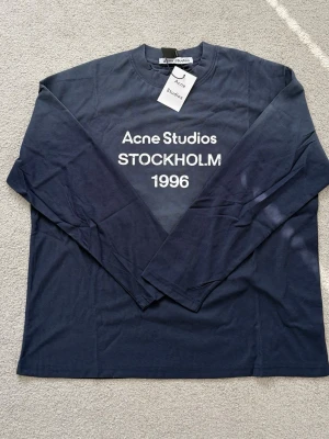 Mörkblå  topp Acne Studios - Mörkblå långärmad topp från Acne Studios med vit text framtill där det står 'Acne Studios STOCKHOLM 1996'. Klassisk rund halsringning och avslappnad passform. Perfekt för dig som gillar stilrena och trendiga plagg.