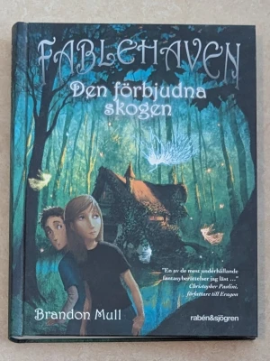 Fablehaven: Den förbjudna skogen - Fantasifylld bok av Brandon Mull med ett mystiskt omslag i gröna och blå toner. Omslaget visar två ungdomar framför en stuga i en magisk skog, omgivna av lysande varelser. Boken ges ut av rabén&sjögren och bjuder på äventyr och magi.