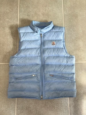 Moncler väst i ljusblå. - NFC Loggan funkar också! Skicka gärna frågor! Snygg ljusblå dunväst från Moncler med hög krage och dragkedja framtill. Västen har två fickor med dragkedja och det klassiska Moncler-logotypen på bröstet. Perfekt för lager-på-lager och ger en clean look!