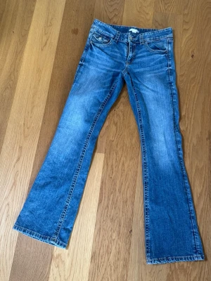 Blå bootcut jeans från Gina Tricot - Snygga blå bootcut jeans från Gina Tricot med klassisk femficksmodell och slitningar för en cool look. Jeansen har normal midja och är tillverkade i mjuk denim med vita kontrastsömmar. Perfekta för dig som gillar en avslappnad men trendig stil. Passar som xs.